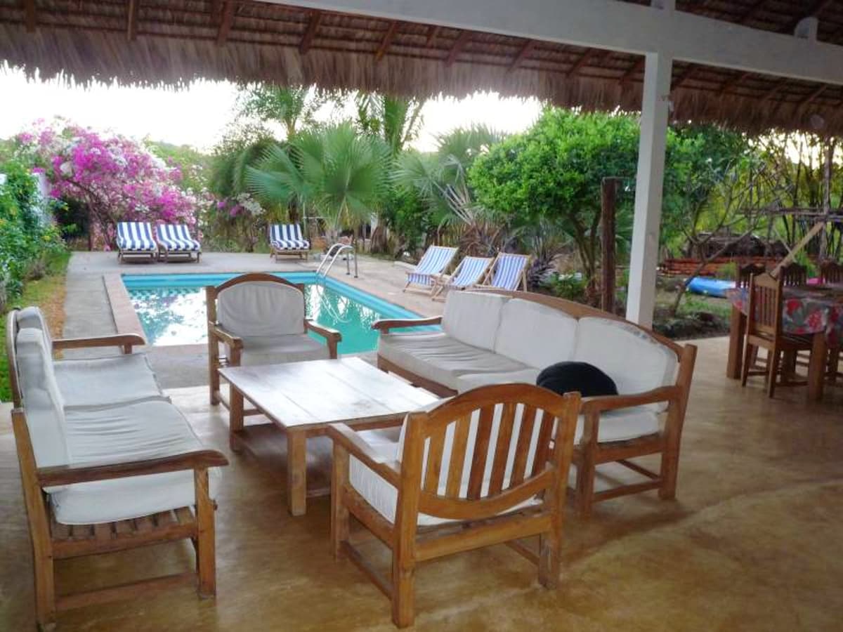 An outdoor seating area features wooden furniture and a coffee table, providing a relaxed space for gatherings. Lush greenery surrounds the area, with a private pool visible in the background and lounge chairs positioned nearby.