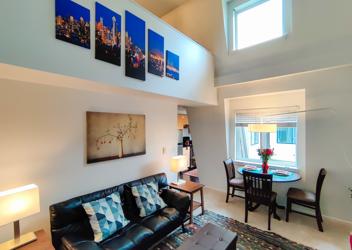 A cozy living area features a black leather sofa adorned with decorative pillows, complemented by a coffee table and illuminated by warm lamps. A dining nook is visible with a round table set for two. Colorful cityscape artwork adorns the wall above.