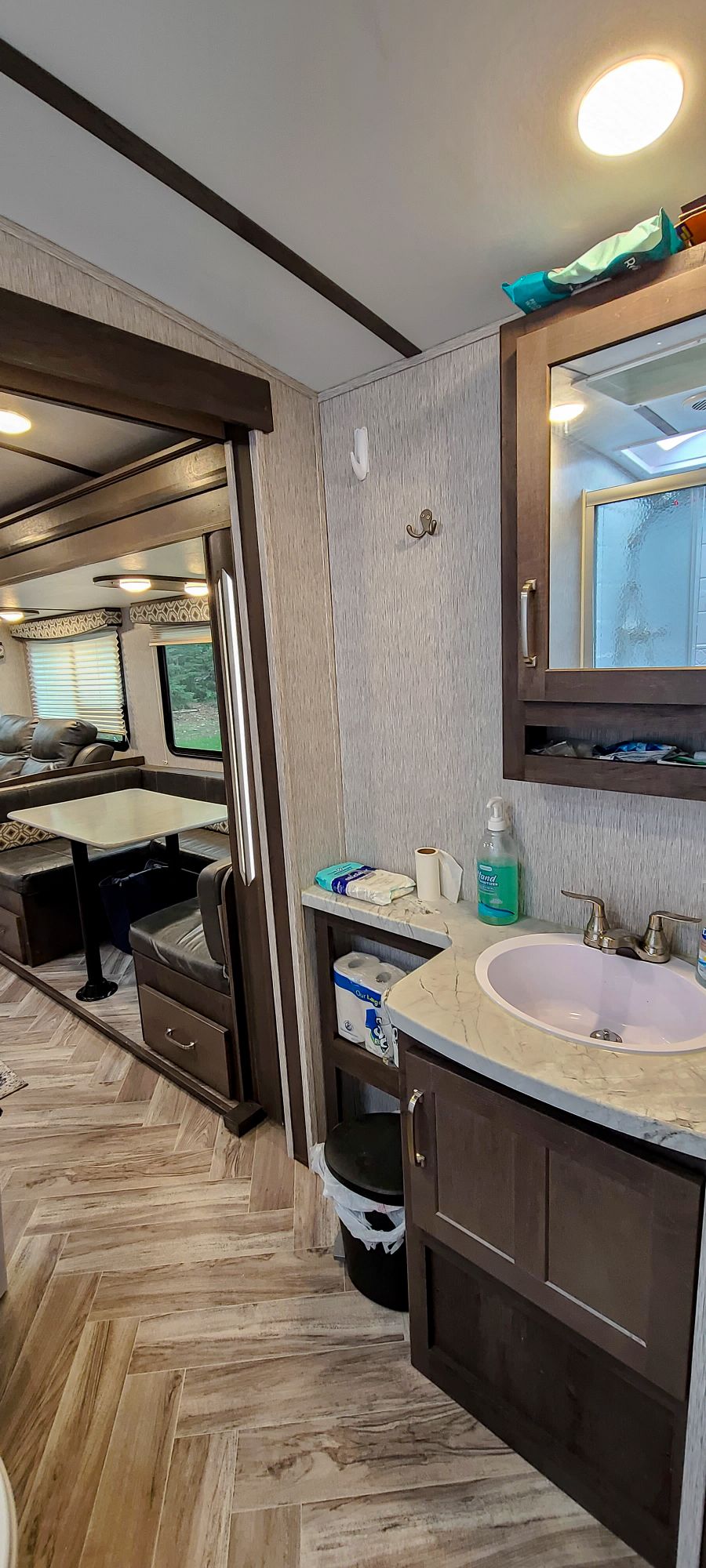 A private bathroom features a compact sink with a light-colored countertop. A tall mirror above reflects the soft indoor lighting. Adjacent to the sink area, wood cabinetry provides storage, and a small space allows for a work station with a table and chairs visible in the background.