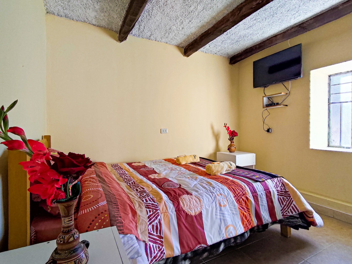 A spacious bedroom features a large bed adorned with a colorful quilt. Decorative flowers are positioned on a bedside table, and a wall-mounted television is visible above a small dresser. Natural light enters through a nearby window framed by exposed wooden beams.