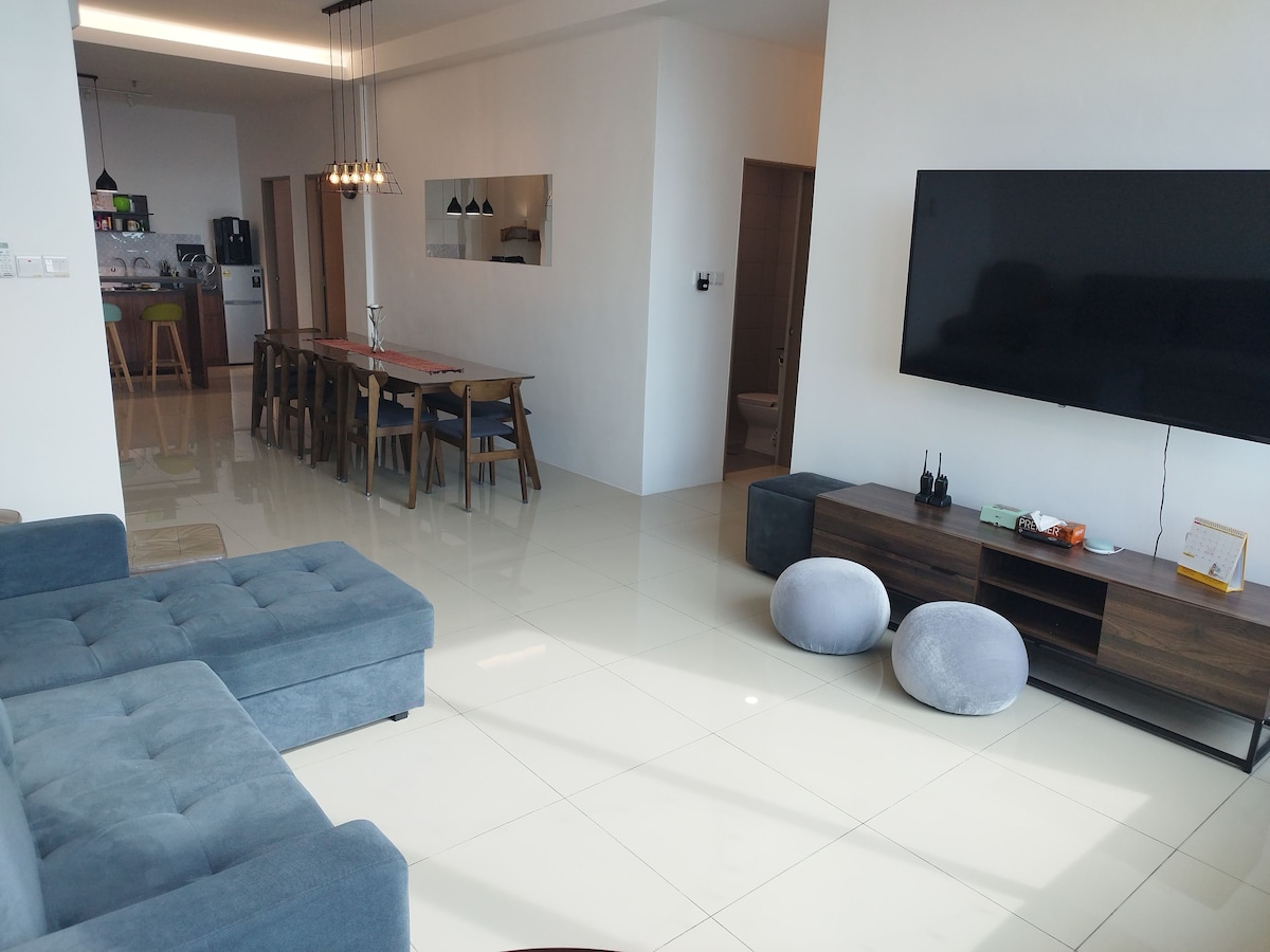 A spacious living and dining area features large windows, allowing natural light to fill the room. A comfortable sectional sofa is paired with two round ottomans. The dining table, designed for ten, is centrally positioned. A television is mounted on the wall, enhancing the entertainment setup.