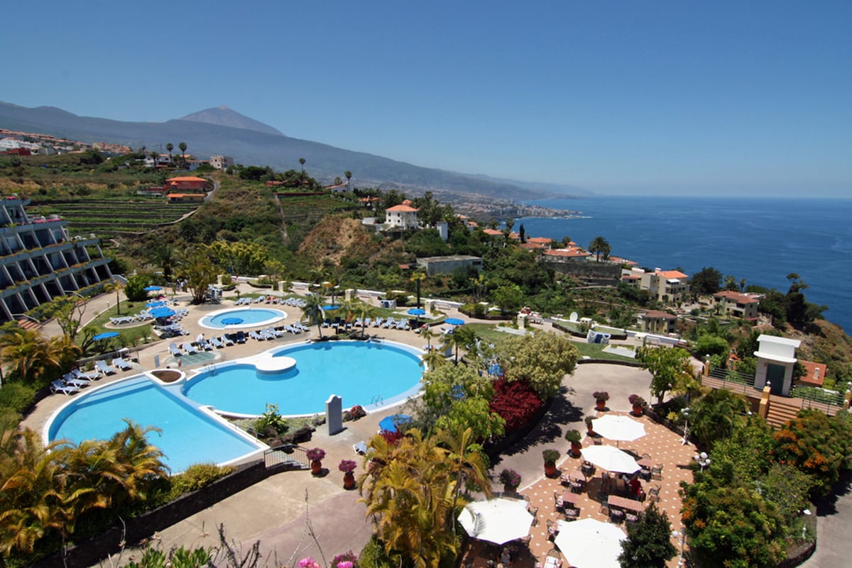 A spacious outdoor area features multiple swimming pools surrounded by lush greenery. Various lounge chairs are available for relaxation, and shaded dining tables provide seating. The serene ocean and mountainous landscape create a scenic backdrop under clear, blue skies.