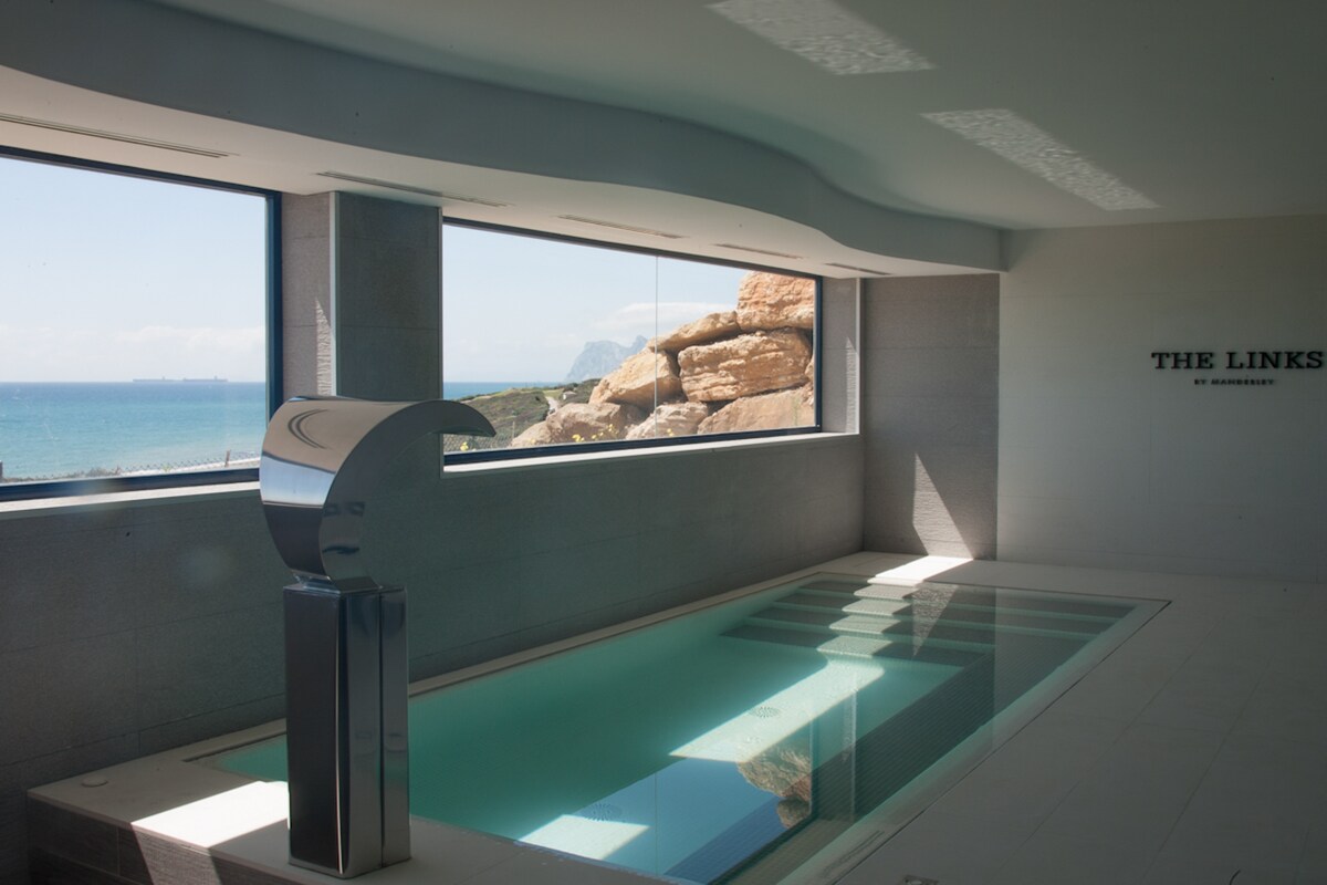 An indoor pool is depicted featuring jet settings and a waterfall. Large glass windows provide views of the sea and rocky landscape. The space is designed with clean lines and neutral tones, emphasizing a relaxed and serene atmosphere.