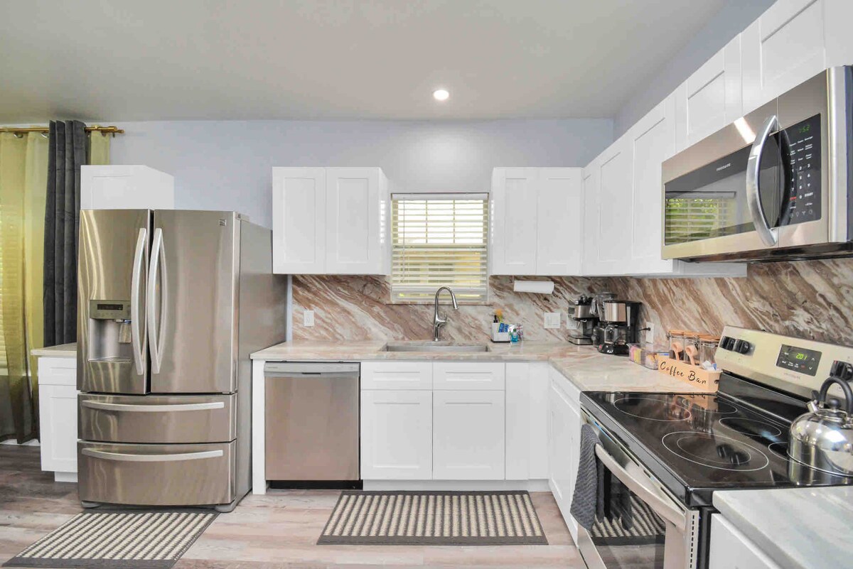 The kitchen space features modern appliances, including a stainless steel refrigerator, dishwasher, and oven. White cabinetry complements the marble backsplash. A window allows natural light to enhance the area, while two area rugs add warmth to the floor.