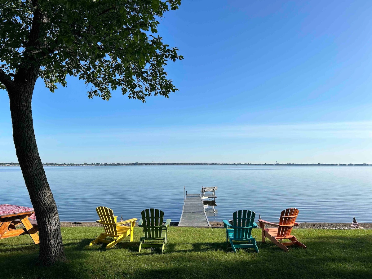 A serene lakeside view features colorful adirondack chairs arranged on grassy land, facing a wooden dock extending into the calm water. A tree provides shade, and the clear blue sky reflects the tranquility of the scene.