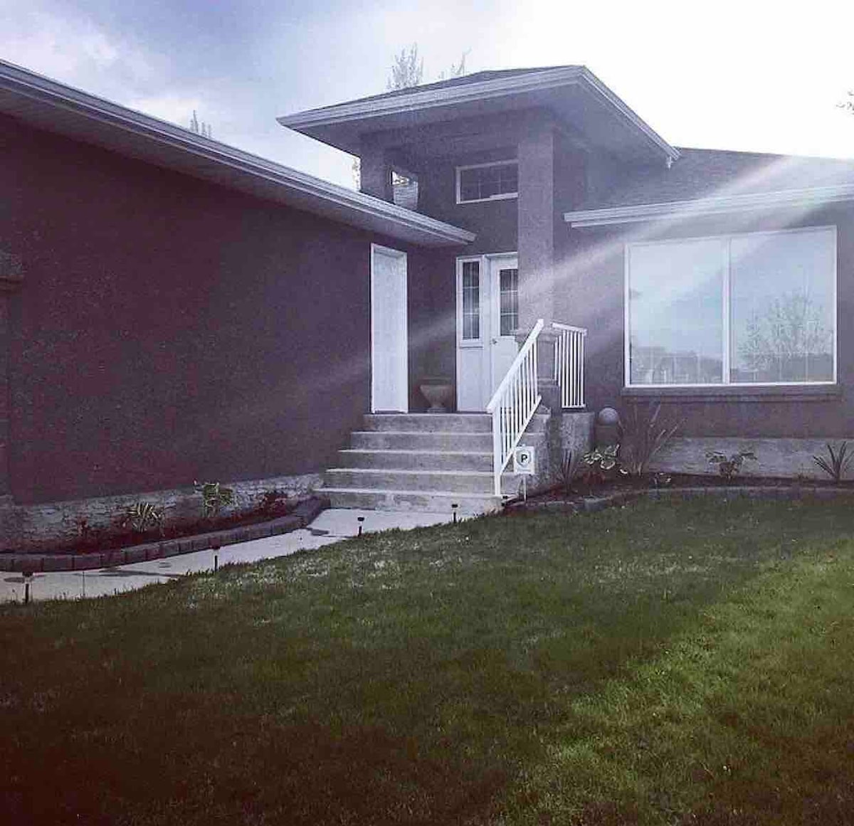 A modern home with a simple façade is presented, featuring a welcoming front entrance with steps leading up to it. Large windows allow natural light to enter, illuminating the inviting entryway. A well-maintained lawn surrounds the house, contributing to a serene outdoor setting.