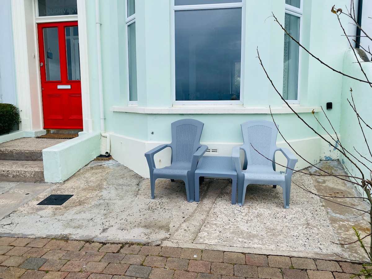 Two light grey lounge chairs are positioned on a stone patio, facing a large window. The entrance features a bright red door surrounded by a soft green exterior. The area is peaceful, perfect for enjoying the sea views.