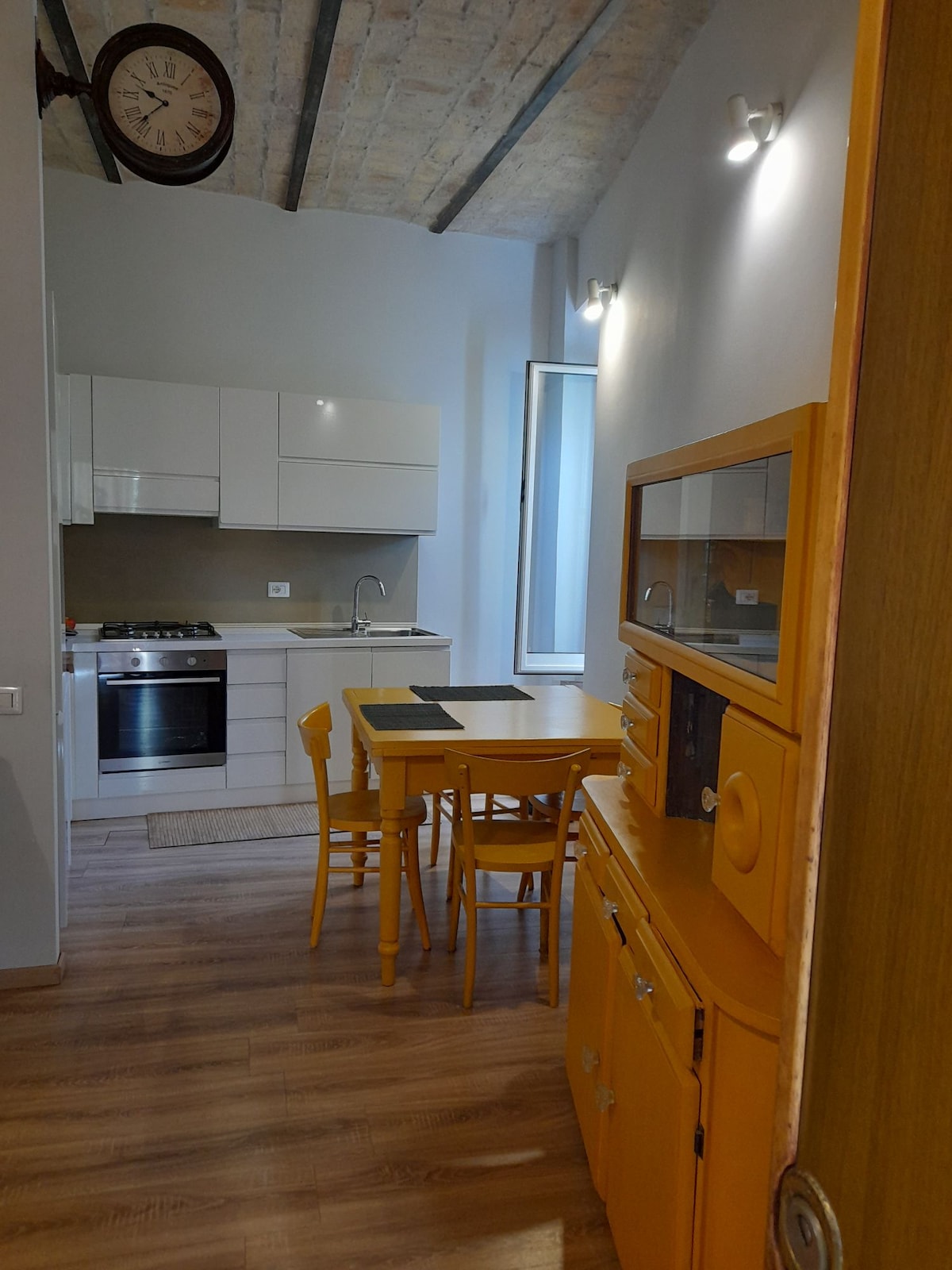 A bright kitchen area is presented, featuring sleek white cabinetry and modern appliances. A small dining table with four yellow chairs is positioned near a window, allowing natural light to fill the space. Wooden flooring and exposed beams add character to the room.