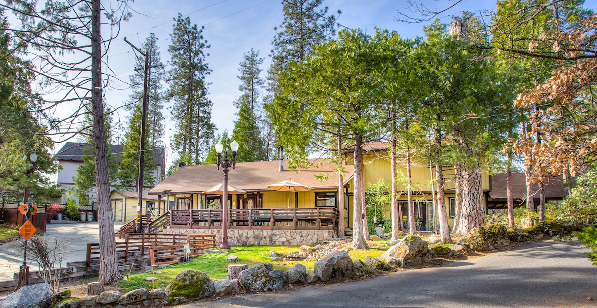 The exterior of the house is visible, featuring a spacious front deck surrounded by tall pine trees. The structure is painted in warm hues, with outdoor seating arrangements on the deck. A gravel path leads to the entrance, showcasing a blend of nature and architecture.