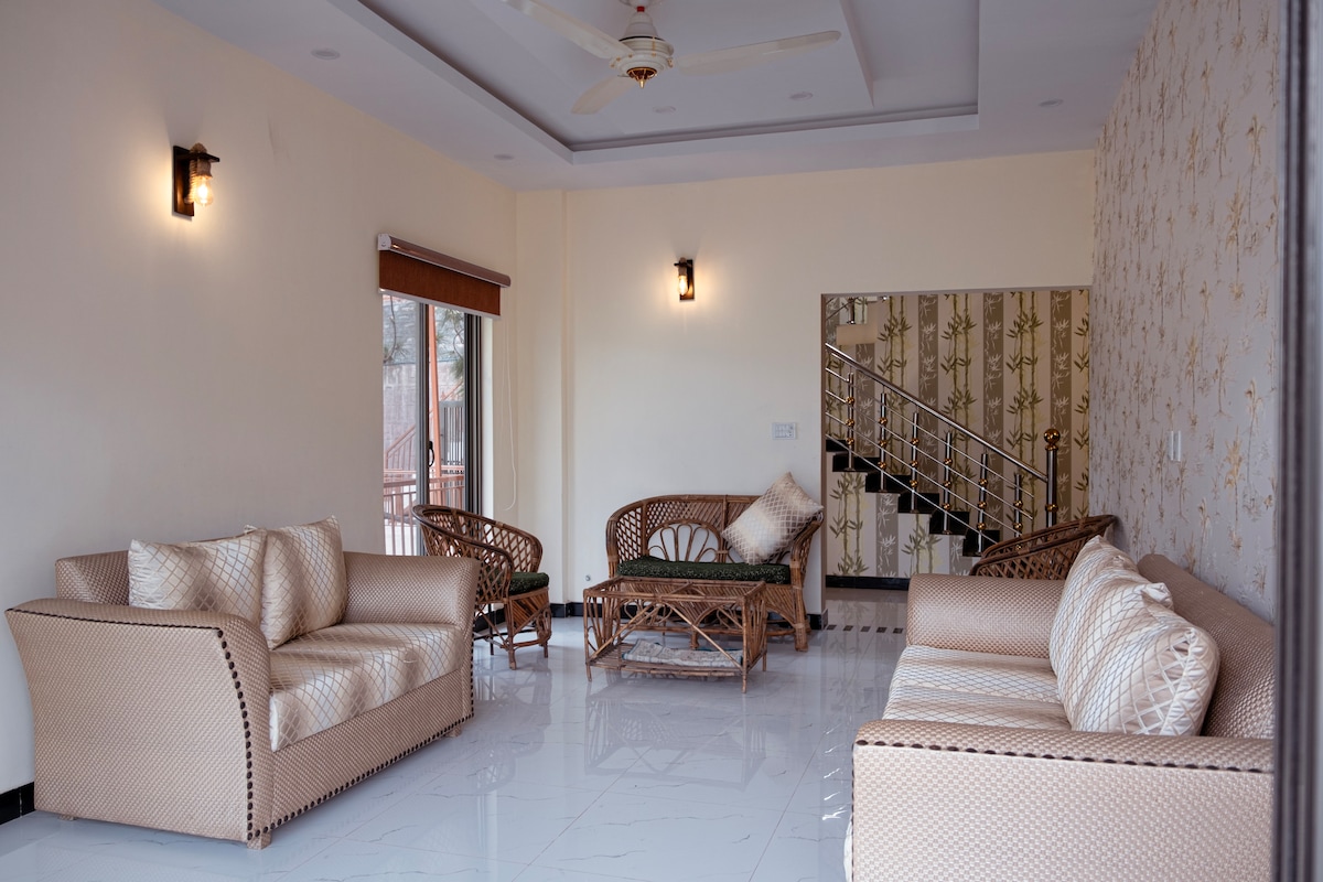 A spacious living area features light-colored seating arrangements that include two sofas and a central coffee table. Natural light enters through large windows, and a staircase is visible in the background, enhancing accessibility. The walls display a subtle pattern that complements the décor.
