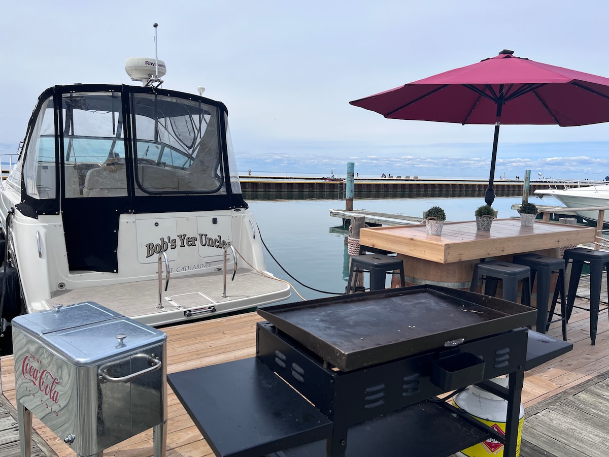 A boat is docked at the marina, featuring a covered cockpit and a name displayed on the stern. A large umbrella shades a dining area with a wooden table and small potted plants. A grill is positioned nearby on a wooden deck, adding to the outdoor space.