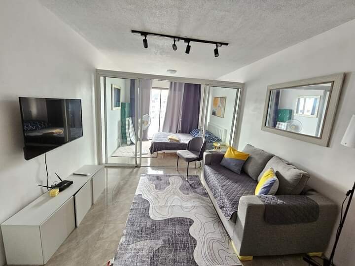 A modern living space features a gray sofa with yellow accents, positioned in front of a television mounted on the wall. Floor-to-ceiling glass doors lead to a bedroom, bringing natural light into the room. A large area rug adds texture to the polished floor.