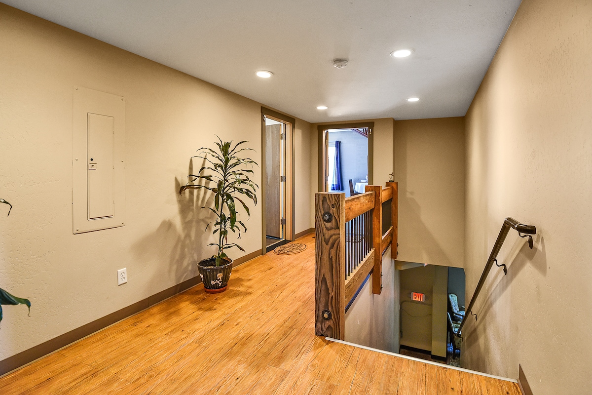 A well-lit hallway features warm wood flooring and neutral walls. A potted plant adds a touch of greenery, while a wooden railing guides the way. Doorways lead to individual rooms, with one visible at the end of the corridor.