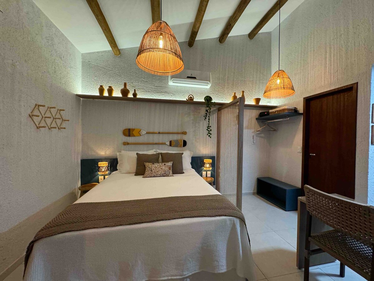 A cozy sleeping area features a spacious bed adorned with neutral linens and a decorative pillow. Woven pendant lights hang from the ceiling, illuminating the room. Shelves with decorative items and wooden paddles add rustic charm, while a closet and seating area provide functionality.