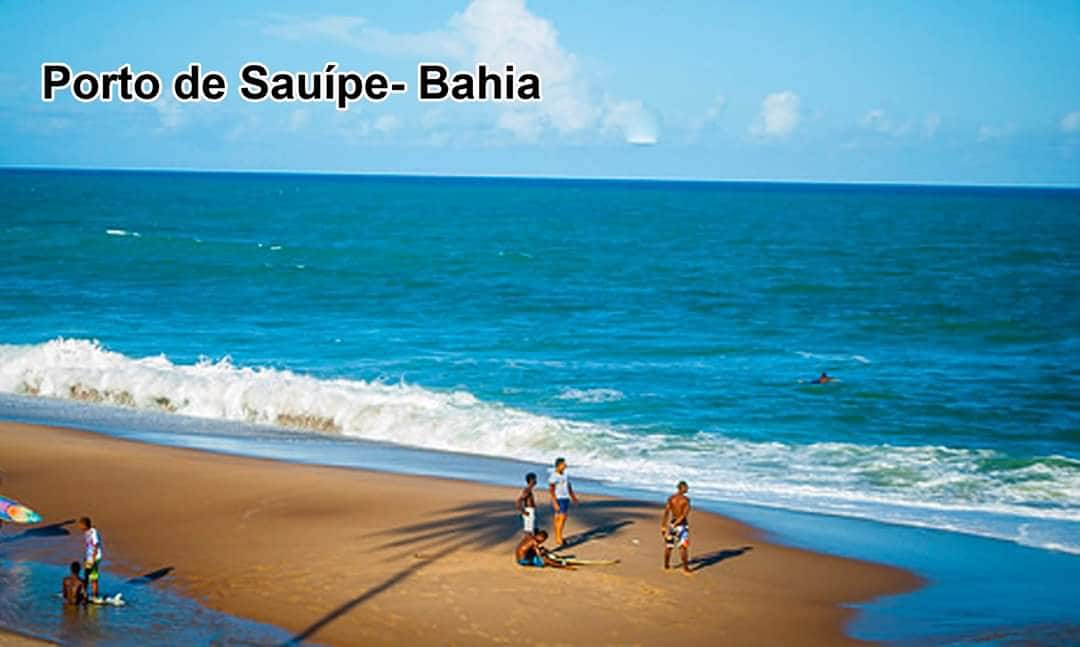A sandy beach extends under a clear blue sky, with gentle waves lapping at the shore. A group of people is engaged in various activities along the water's edge, while colorful umbrellas dot the area, providing shade.
