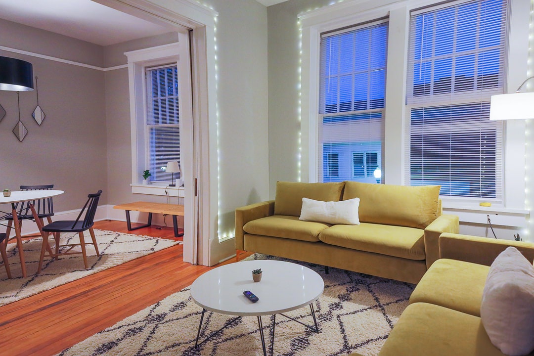 A bright living area features large windows adorned with soft blinds, allowing natural light to fill the space. A yellow couch sits beside a round coffee table. A dining area is visible, with a small circular table and four chairs. Warm ambient lighting enhances the inviting atmosphere.