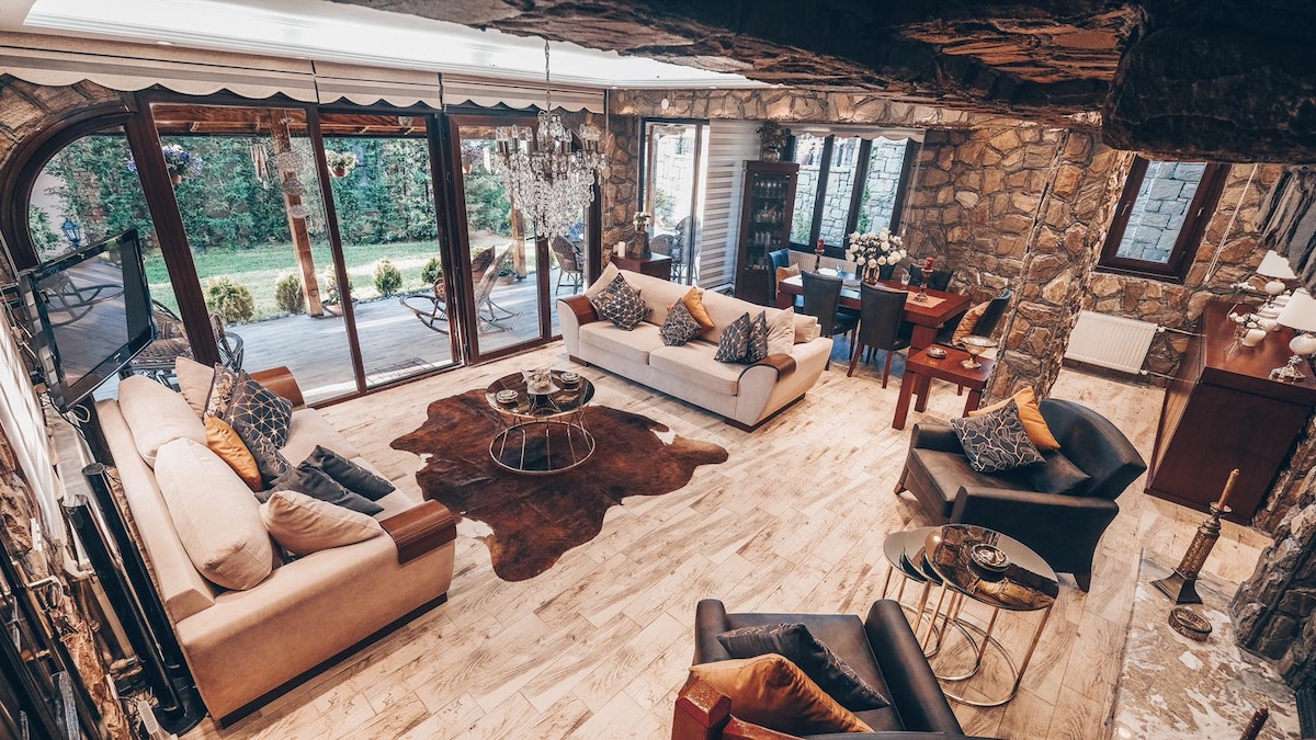 The spacious living area features natural stone walls complemented by wooden accents. Comfortable seating includes two cream sofas and stylish black chairs. A contemporary coffee table sits at the center, surrounded by decorative cushions. Large glass doors open to a lush garden, enhancing the room's ambiance.