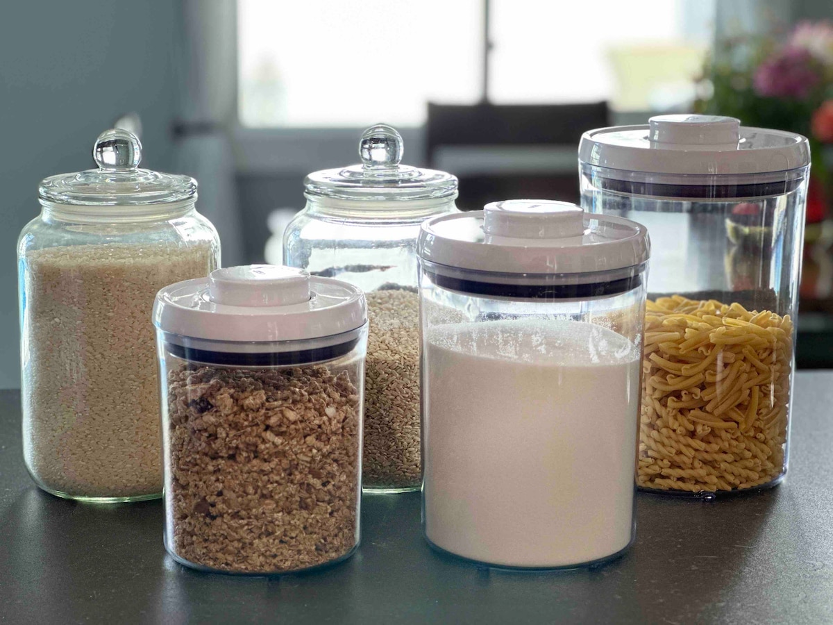 Various glass storage containers filled with ingredients are displayed on a kitchen countertop. The containers include rice, oats, sugar, and pasta. Each has a clear lid, allowing for easy visibility of contents. Natural light brightens the space, enhancing the organized appearance.