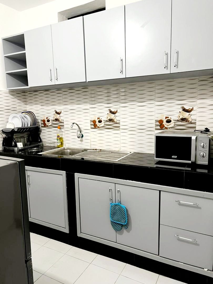 A modern kitchen displays sleek gray cabinetry and a black countertop. Wall tiles with a wavy design provide a textured backdrop. A stainless steel sink is positioned between essential kitchen appliances, including a microwave and a coffee maker. Dishware is neatly arranged on open shelves above.