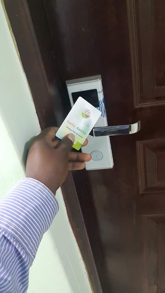 A hand is shown holding an electronic key card, positioned near the door lock of a wooden door. The door features a sleek handle and a panel for card access, emphasizing the modern security system of the accommodation.