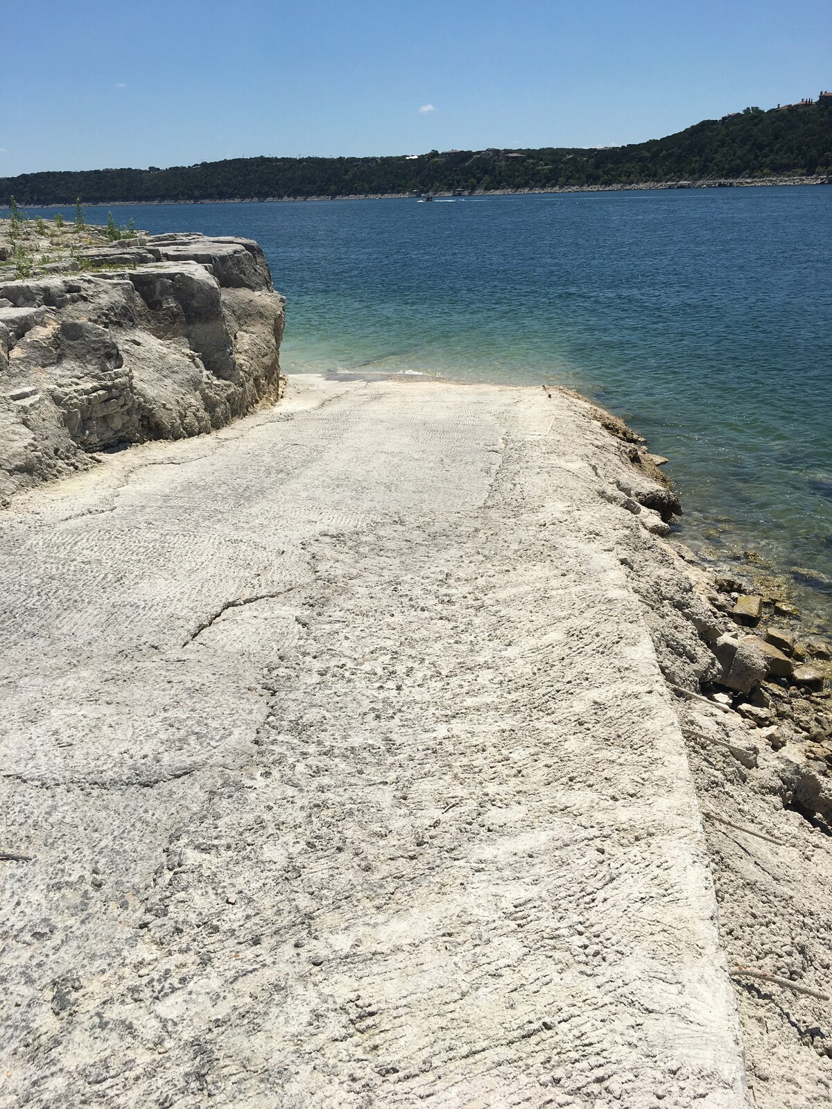 A sunlit shoreline features smooth rock ledges leading to the clear waters of Lake Travis. The water reflects shades of blue and green, providing a refreshing spot for lounging or swimming. A gently sloping edge allows for easy access to the water, surrounded by natural scenery.