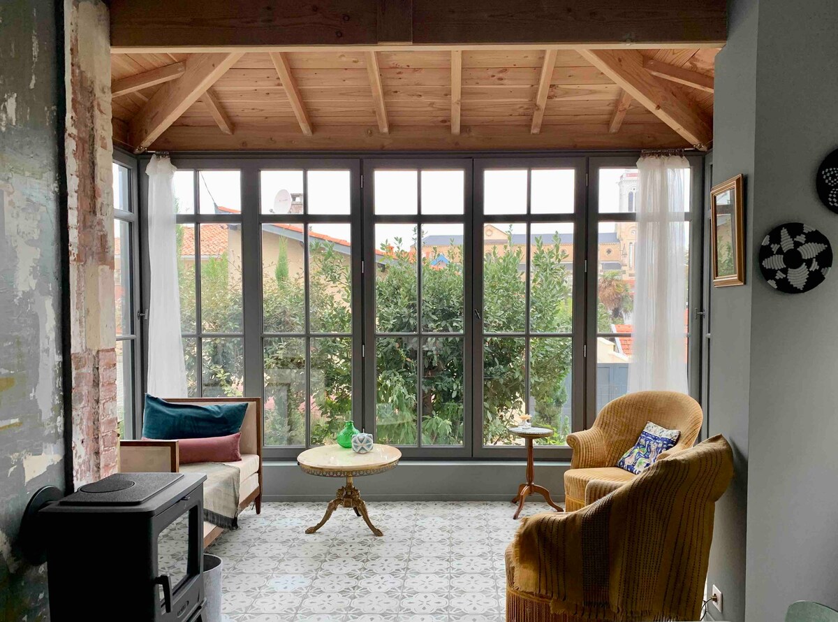 A sunlit sitting area features large windows framing views of greenery. Comfortable seating, including a plush yellow chair and a sofa, surrounds a round coffee table. The room showcases a combination of rustic and contemporary elements with exposed beams and patterned tiles.