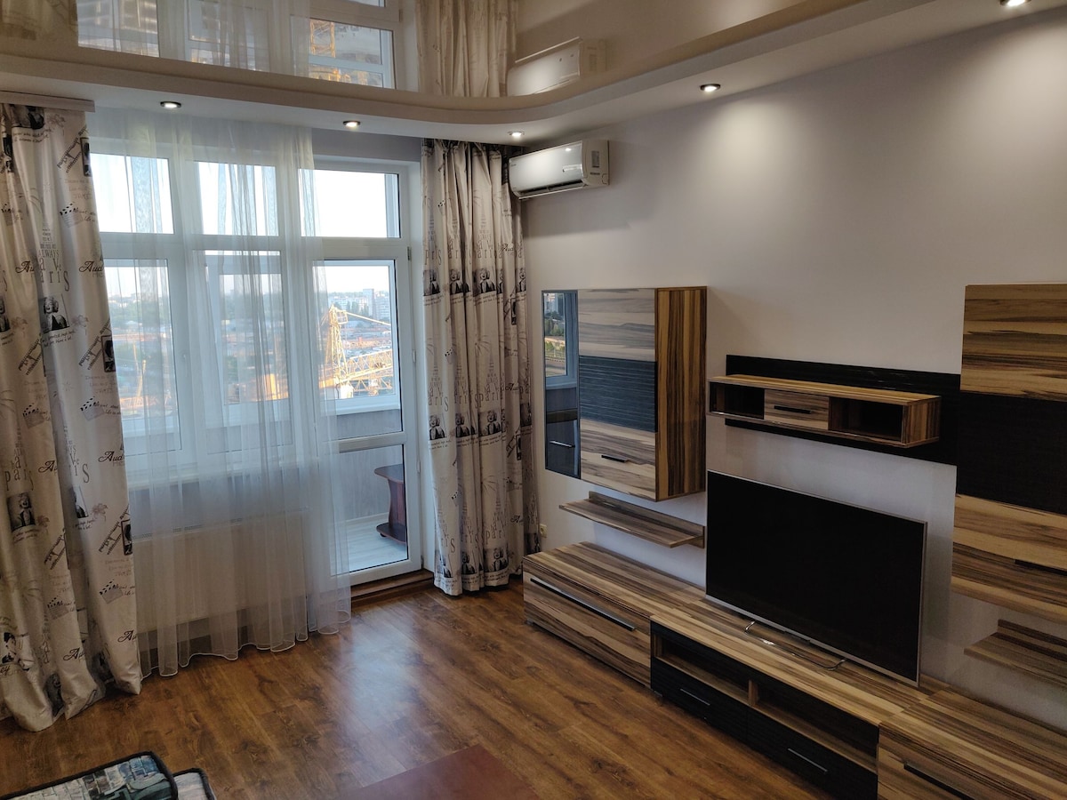 A well-lit living area features large windows with sheer curtains allowing natural light to filter in. Stylish wooden furniture includes a media console with a television and decorative shelving. The warm-toned wooden flooring complements the contemporary design of the space.