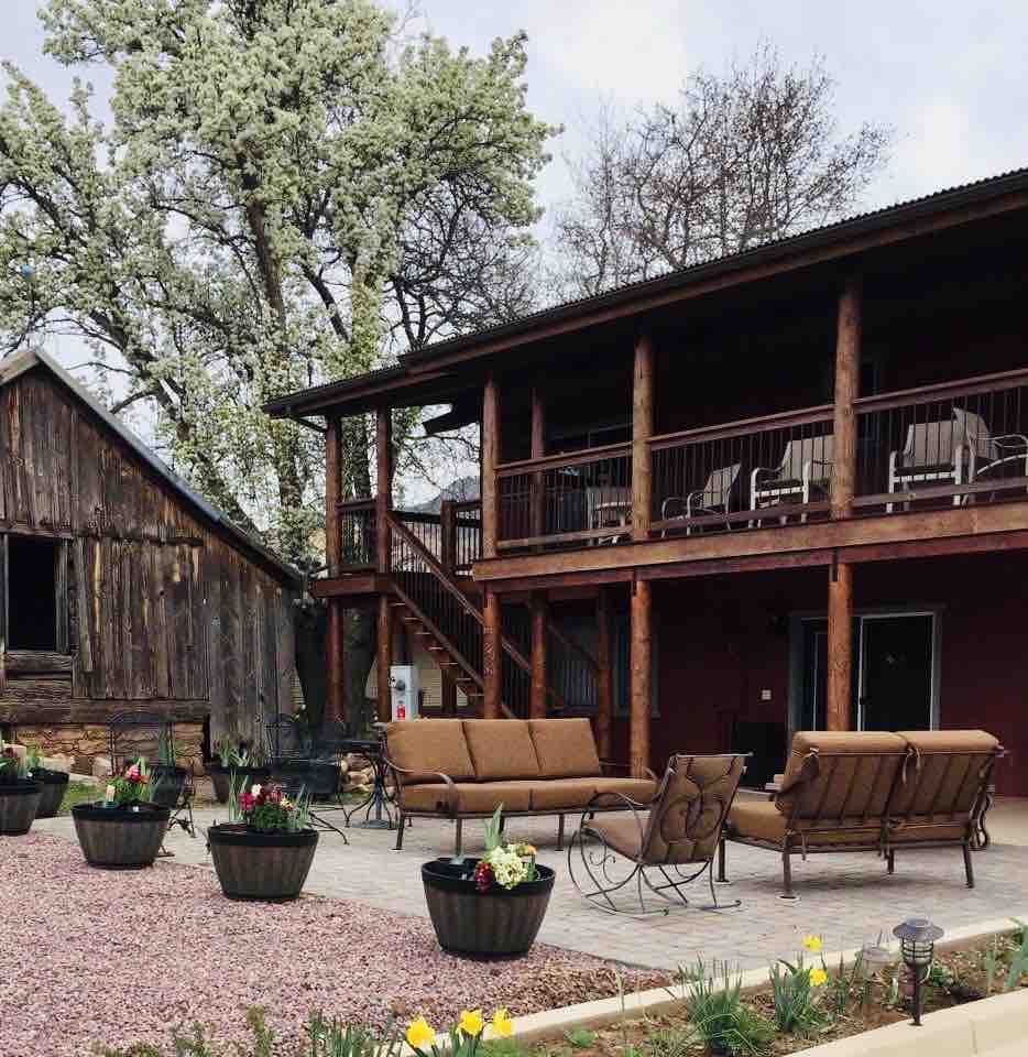 A spacious outdoor seating area features a suite of brown lounge chairs and a sofa, positioned on a stone patio surrounded by blooming flowers. The upper deck provides access via a staircase, enhancing outdoor gathering possibilities, all set against a rustic wooden structure.