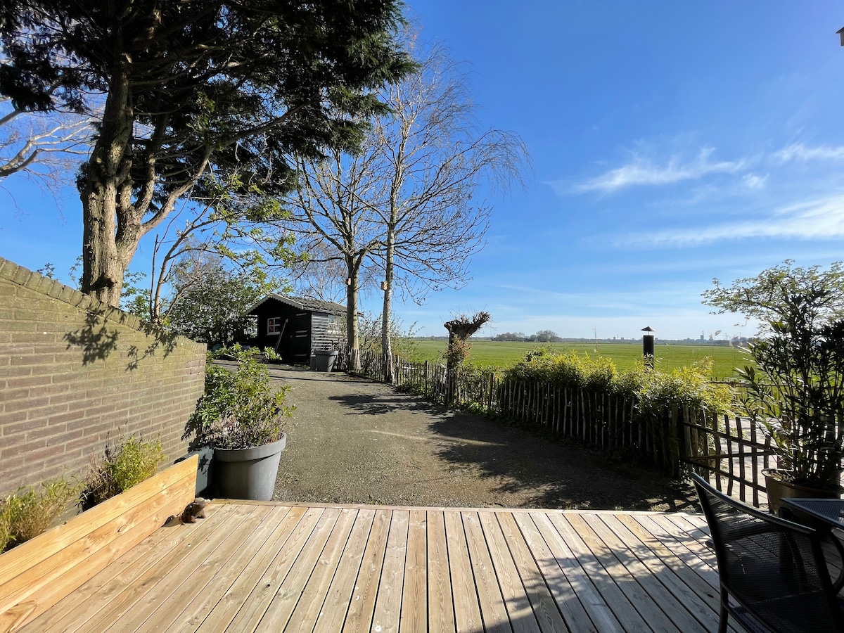 A wooden terrace features space for outdoor seating, overlooking lush green fields under a clear blue sky. Nearby trees provide shade, while a rustic cabin stands in the distance, framed by the natural landscape.