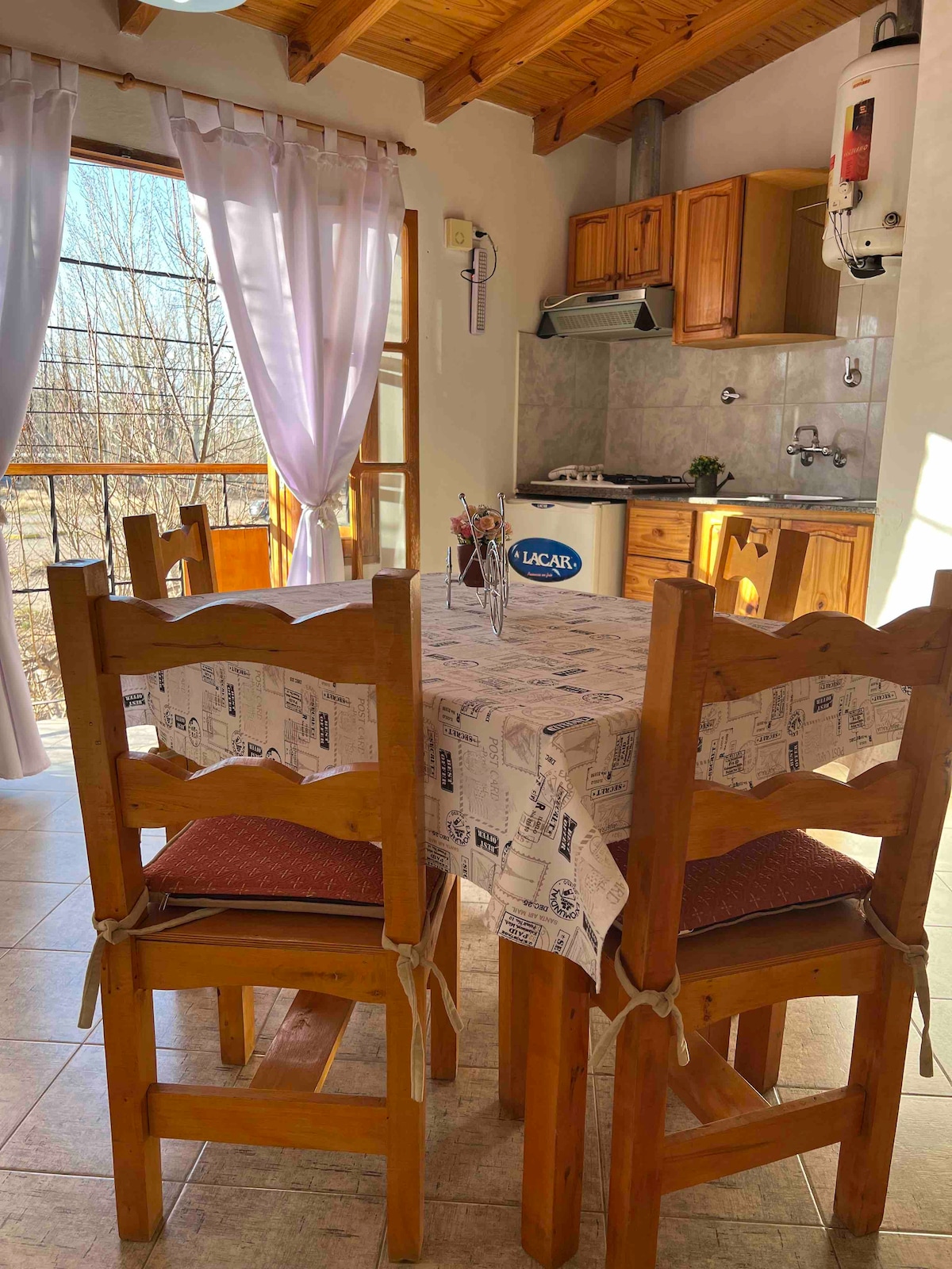 A cozy dining area features a wooden table surrounded by four chairs. Natural light fills the space through large windows, partially draped with light curtains. A kitchenette with wooden cabinetry is visible in the background, complemented by neutral-toned walls and tiled flooring.