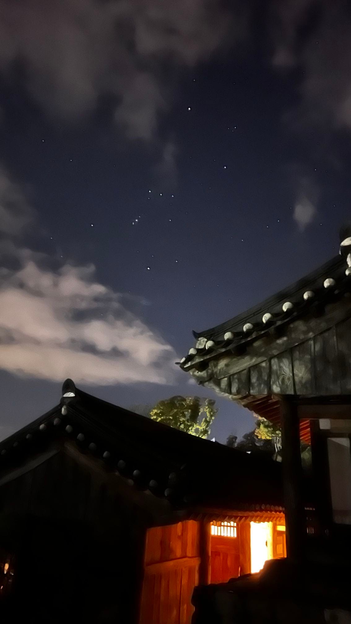 The image captures the night sky above traditional architecture, featuring ornate rooflines with decorative eaves. Stars are visible against a dark blue background, complemented by wispy clouds. The warm glow of light from the building hints at the inviting interior.