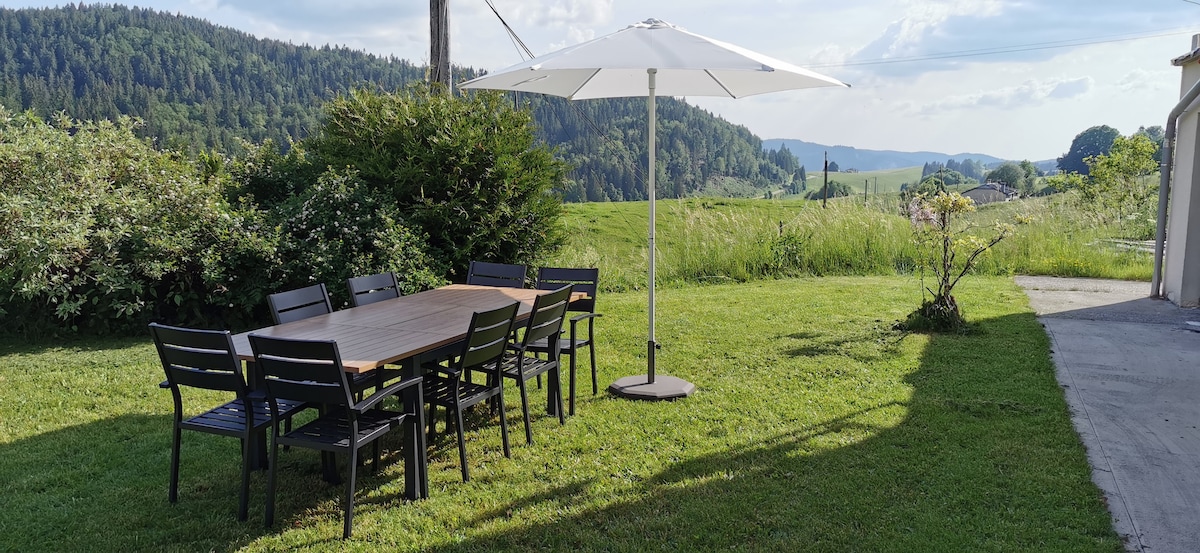 An outdoor dining area is situated on a grassy lawn, featuring a large wooden table surrounded by black chairs. A sizable umbrella provides shade, with rolling green hills visible in the background, enhancing the tranquil setting.
