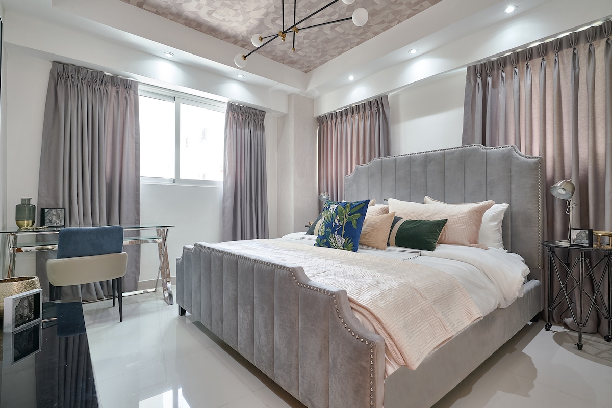 A modern bedroom features a spacious bed with light-colored bedding and decorative cushions. Soft, neutral tones are complemented by dark curtains. A work desk with a chair is positioned near a window, allowing natural light to illuminate the room. Decorative accents are found throughout the space.