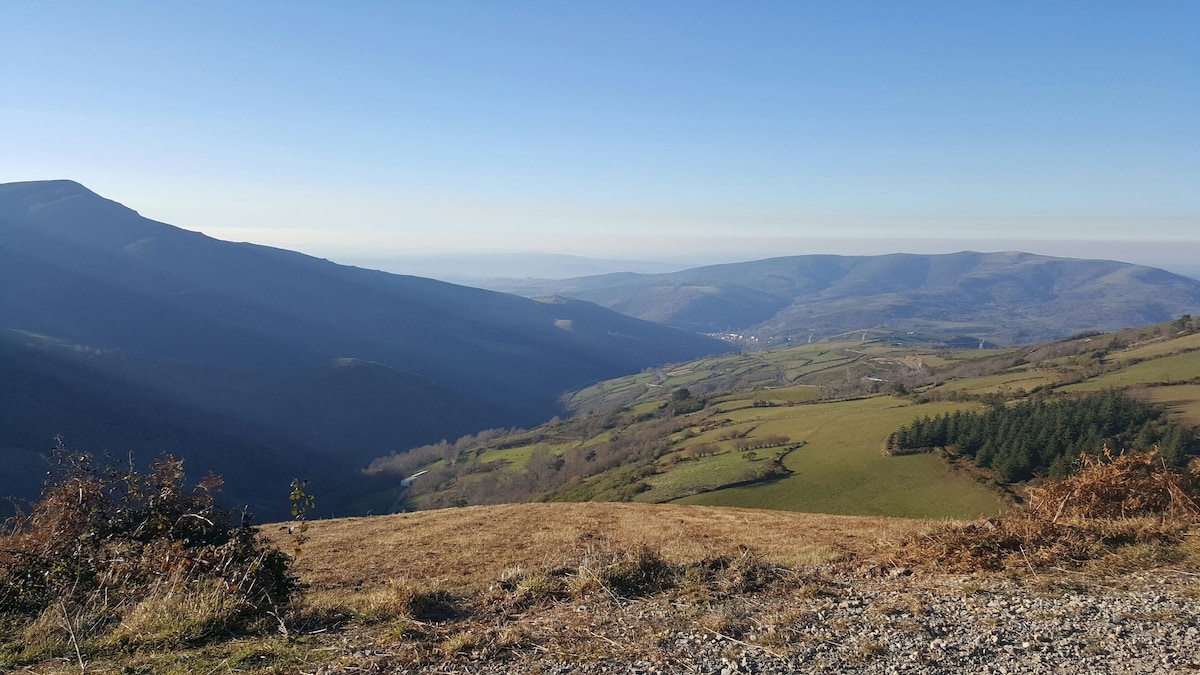 A panoramic view showcases rolling hills and valleys, creating a serene landscape under a clear blue sky. Vegetation varies across green pastures and distant mountains, accentuating the natural beauty of the surrounding area.
