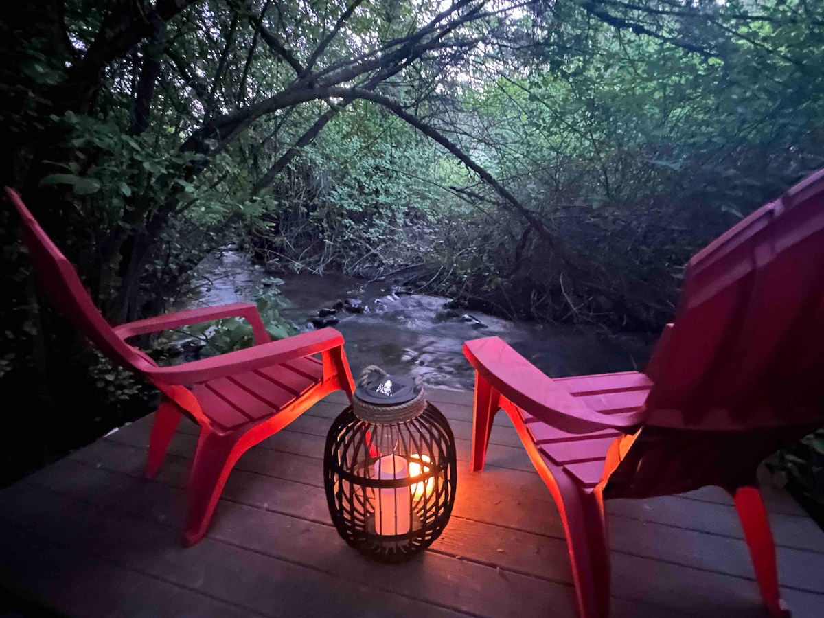 Two red chairs are positioned on a wooden deck beside a gently flowing creek, surrounded by lush greenery. A lantern with a warm glow rests between the chairs, creating a serene ambiance as twilight descends.