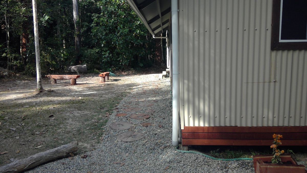 A gravel pathway leads from the corner of a corrugated metal structure, surrounded by lush green foliage. Two stone seating areas with wooden benches are visible nearby, providing a place to relax and enjoy the natural surroundings.