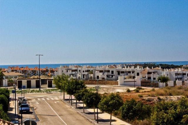 A view of a quiet residential area features well-maintained white buildings and a wide road lined with trees. The distant blue sea is visible, providing a serene backdrop, while clear skies enhance the overall tranquility of the setting.