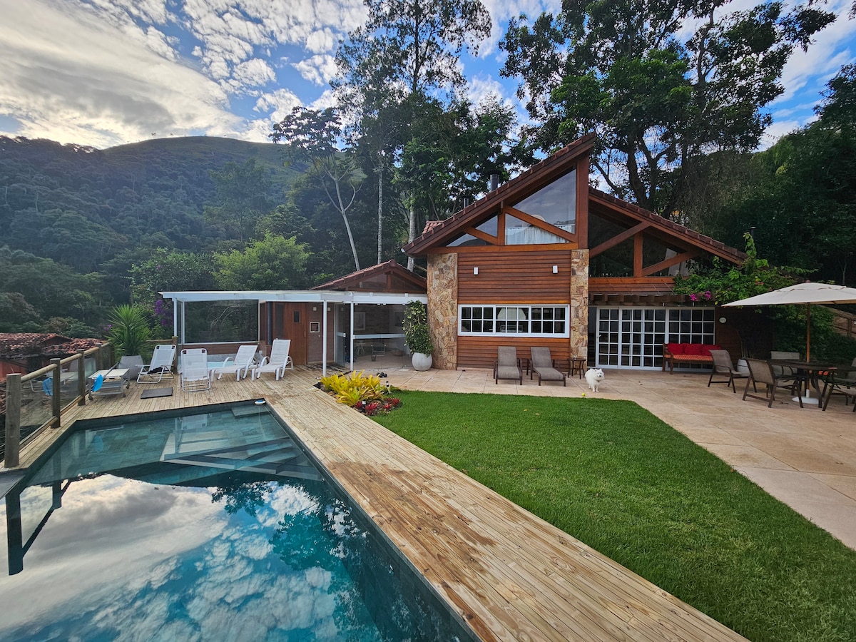 A spacious outdoor area features a wooden deck surrounding a pool. The villa, characterized by its wooden structure and large windows, is set against a backdrop of lush greenery. Sun loungers and dining furniture are arranged nearby, providing options for relaxation and dining.
