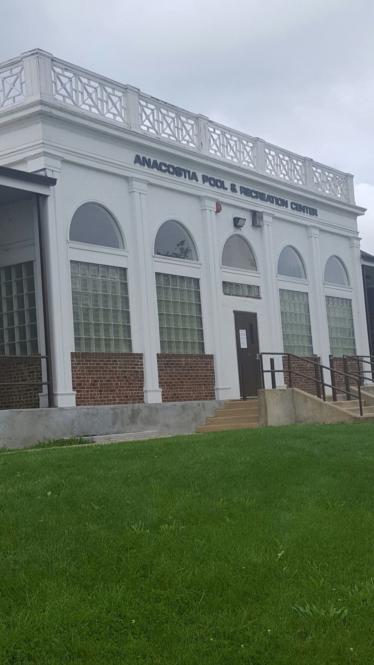 The exterior of the Anacostia Pool and Recreation Center is displayed, featuring a white façade with large, rounded arch windows. A decorative railing crowns the building, adding architectural interest. The surrounding grassy area is maintained, enhancing the inviting atmosphere of the facility.
