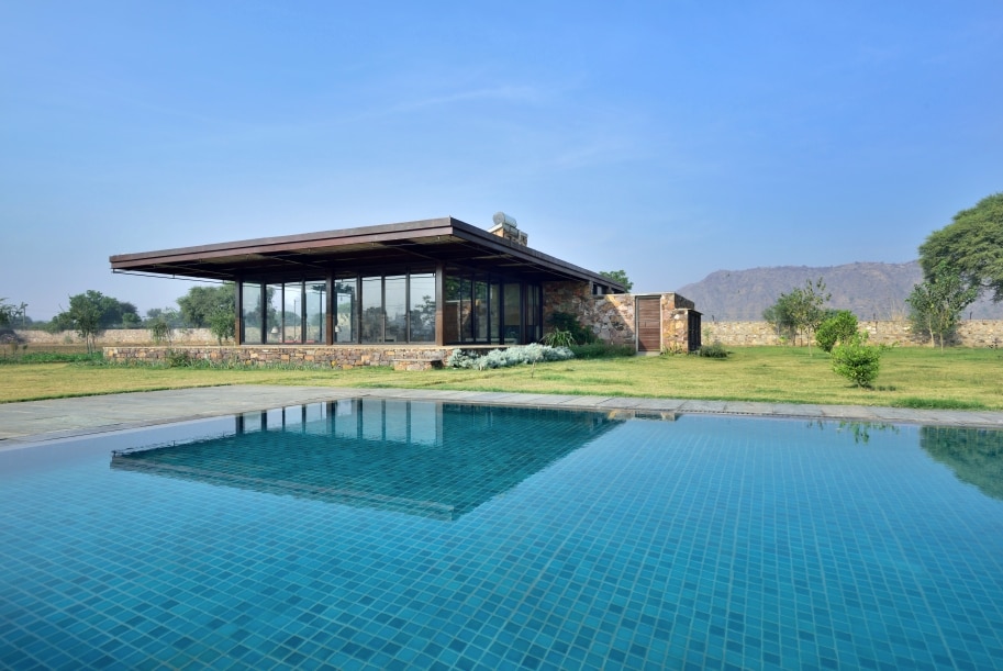 A modern glass house features large windows reflecting the serene landscape. The inviting swimming pool, visible in the foreground, complements the lush green lawn and the distant hills, creating a tranquil retreat atmosphere.