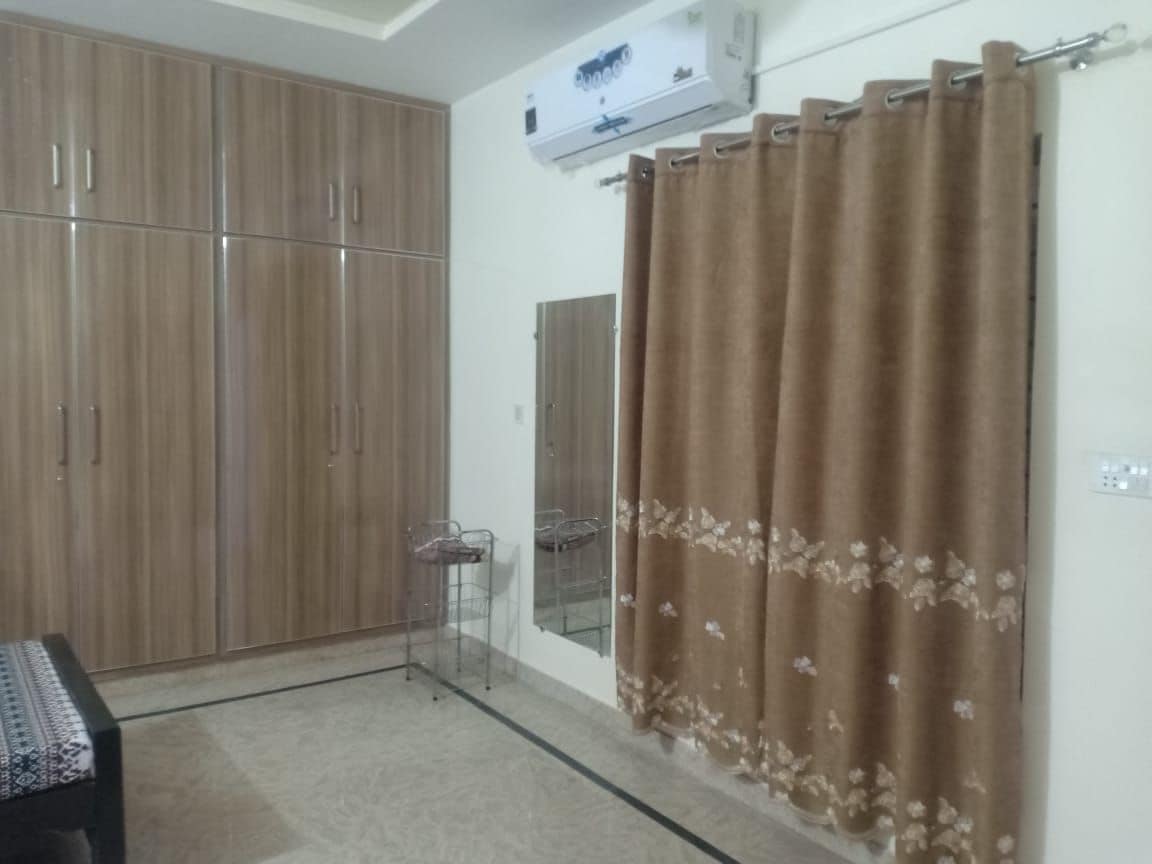 A well-lit room features a large wardrobe with wooden cabinetry, offering ample storage space. A full-length mirror is mounted on the wall, complemented by a curtain in warm tones with floral designs. The floor is tiled, and air conditioning is installed for comfort.