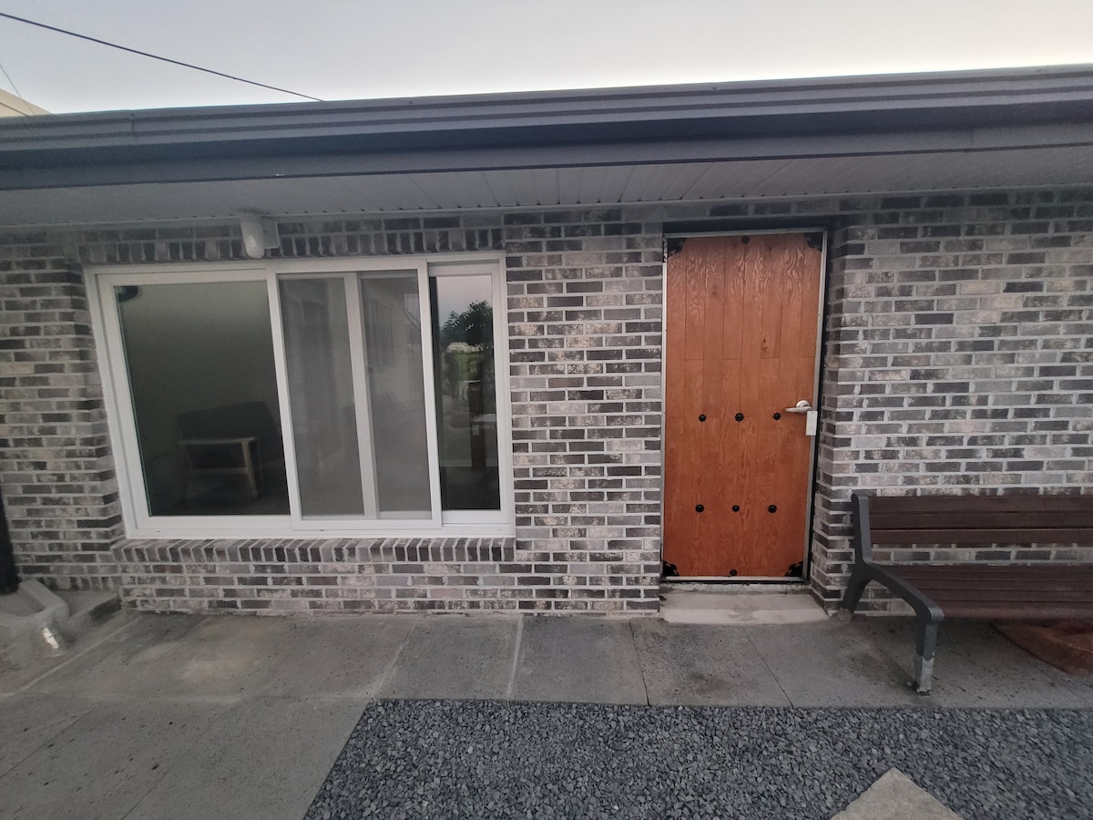 The exterior of a modern gray brick building is visible, featuring a wooden front door with metallic handles. A large glass window next to the door allows natural light into the interior. A small bench is placed beside the entrance.