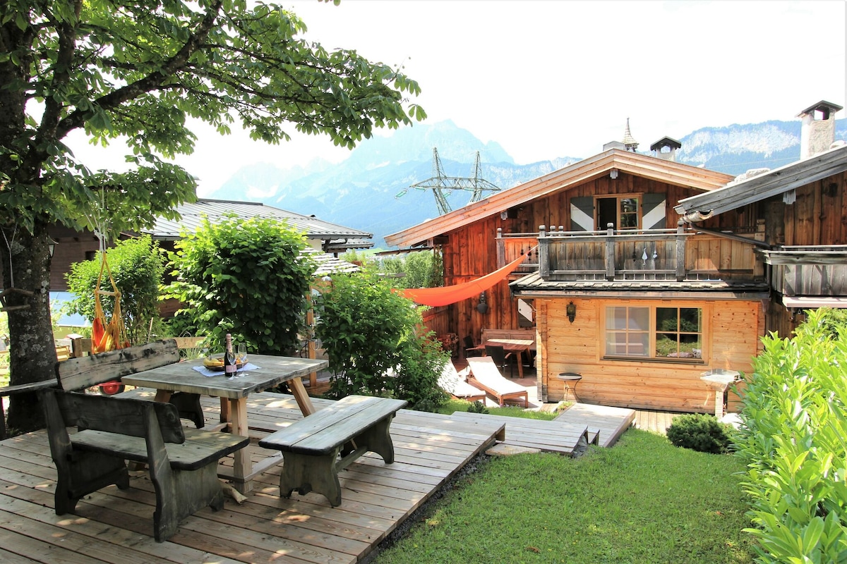An outdoor terrace is visible, featuring rustic wooden picnic tables surrounded by greenery. The chalet's wooden structure and balcony are seen, with a vibrant hammock hung between trees. Breathtaking mountain views rise in the background, enhancing the natural setting.