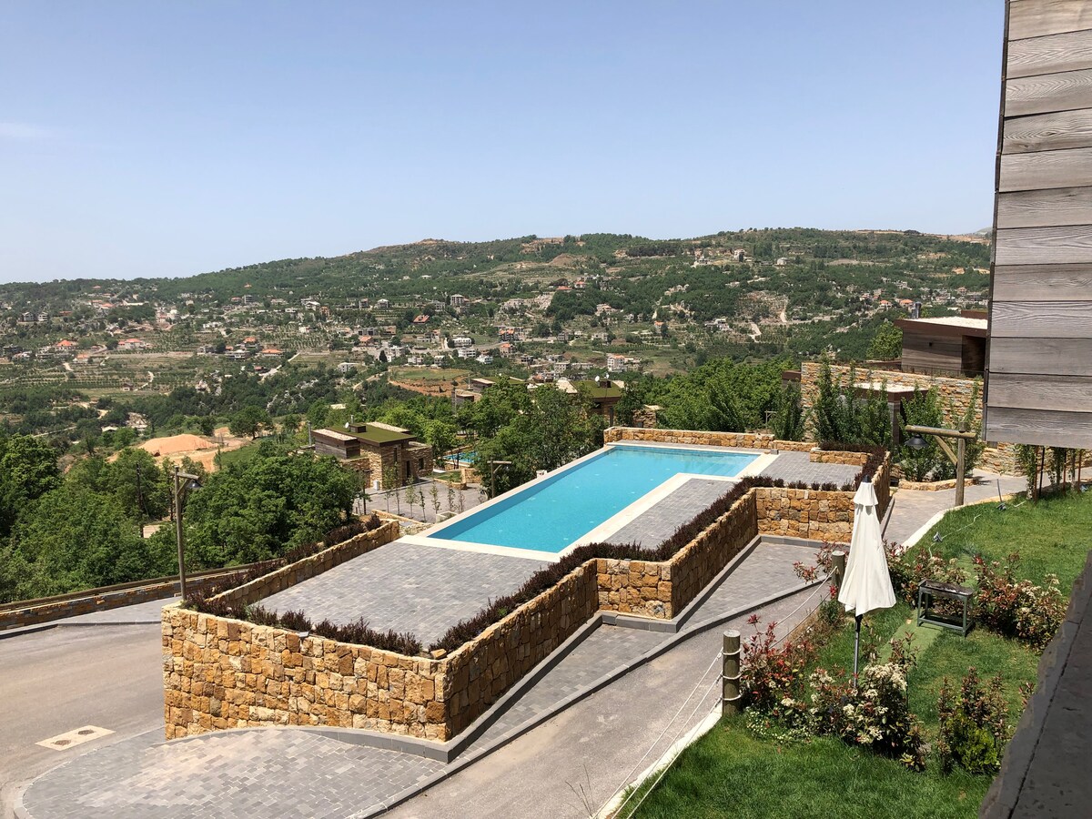 An outdoor swimming pool is situated on a stone terrace, surrounded by landscaped gardens. Rolling hills and distant homes create a scenic backdrop, while an umbrella provides shade nearby. The clear sky enhances the peaceful atmosphere of this elevated location.