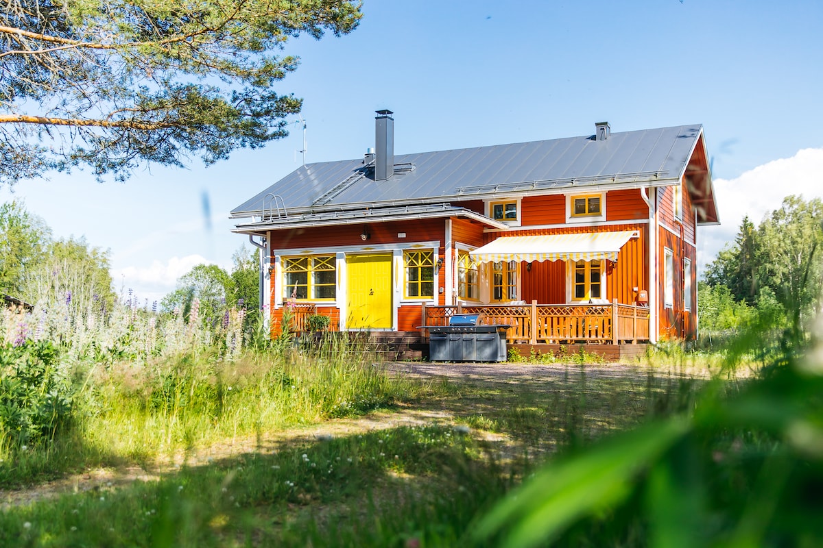 A bright red wooden house is set against a clear blue sky, featuring a welcoming yellow front door. Large windows allow natural light to enhance the exterior, complementing the surrounding greenery. A covered deck provides space for relaxation, with additional outdoor seating visible.