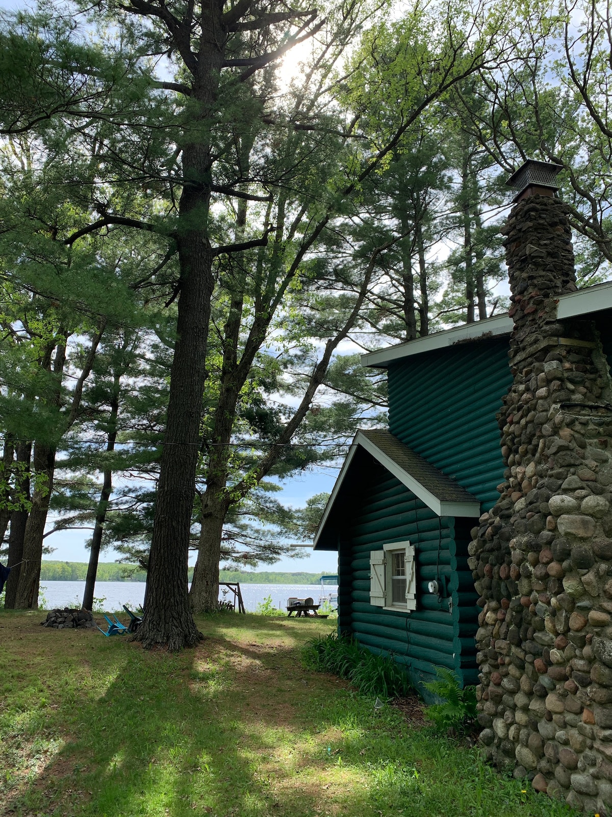 The log cabin features a rustic stone chimney and stands among tall trees, with sunlight filtering through the leaves. A serene view of the lake is visible in the background, offering a peaceful setting for outdoor activities.
