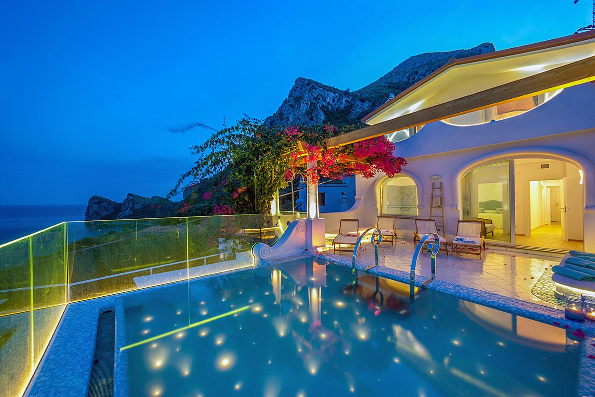 The tranquil scene features a private swimming pool illuminated under the evening sky, reflecting the surrounding landscape. Lush bougainvillea frames the villa, while large glass doors offer views into the open-plan living spaces. The serene atmosphere is enhanced by the coastline in the distance.