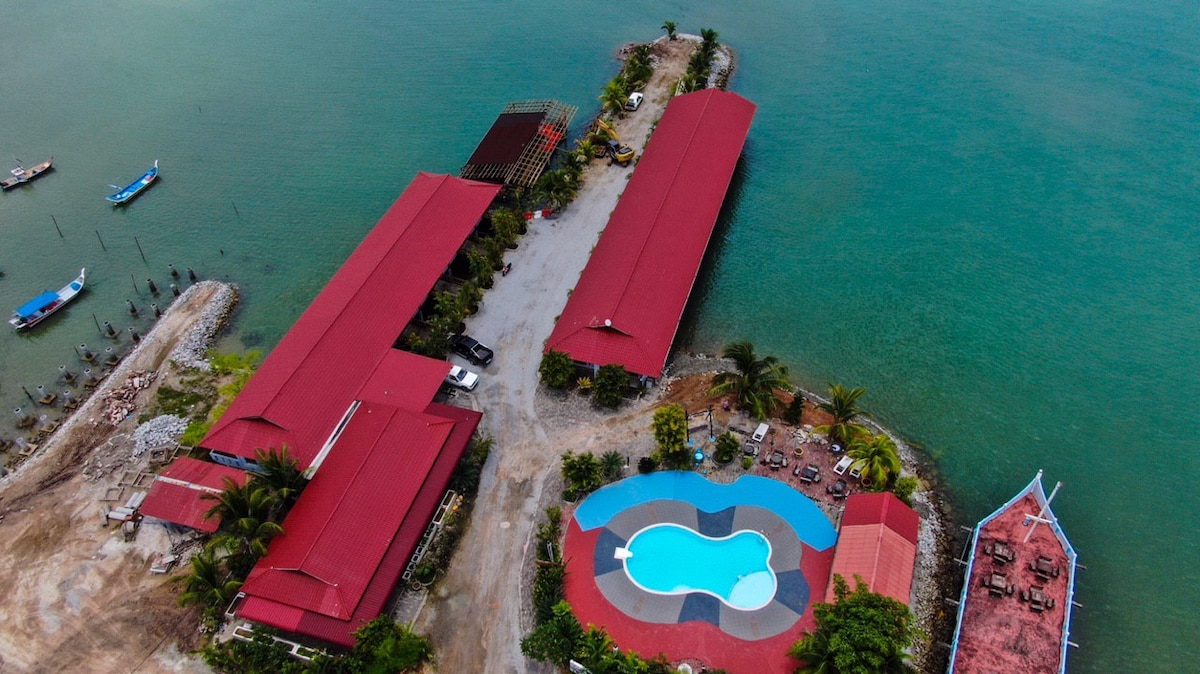 An aerial view showcases the layout of Floating House de Bamboo, highlighting its red-roofed structures surrounded by waters. A swimming pool is visible, featuring a unique shape, with lounge chairs positioned nearby. Boats can be seen floating along the shoreline, enhancing the coastal atmosphere.