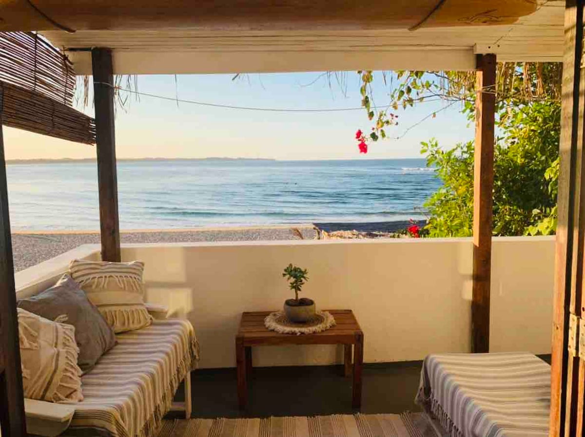 A comfortable outdoor seating area features two striped sofas arranged around a wooden table. Potted plants adorn the table, and vibrant flowers add a touch of color. The view overlooks calm waters and a sandy beach, framed by a clear sky and gentle coastal foliage.