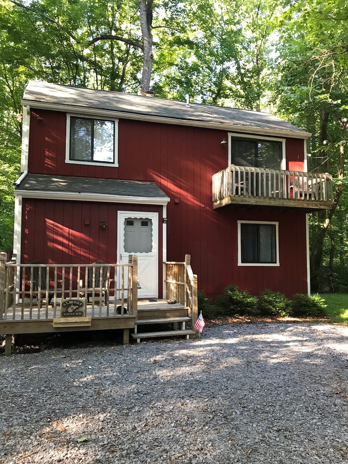 A two-story red cabin is surrounded by lush greenery, featuring a wooden front porch with a railing. A small balcony is visible on the upper level. Gravel leads up to the entrance, and a decorative sign is placed near the porch.